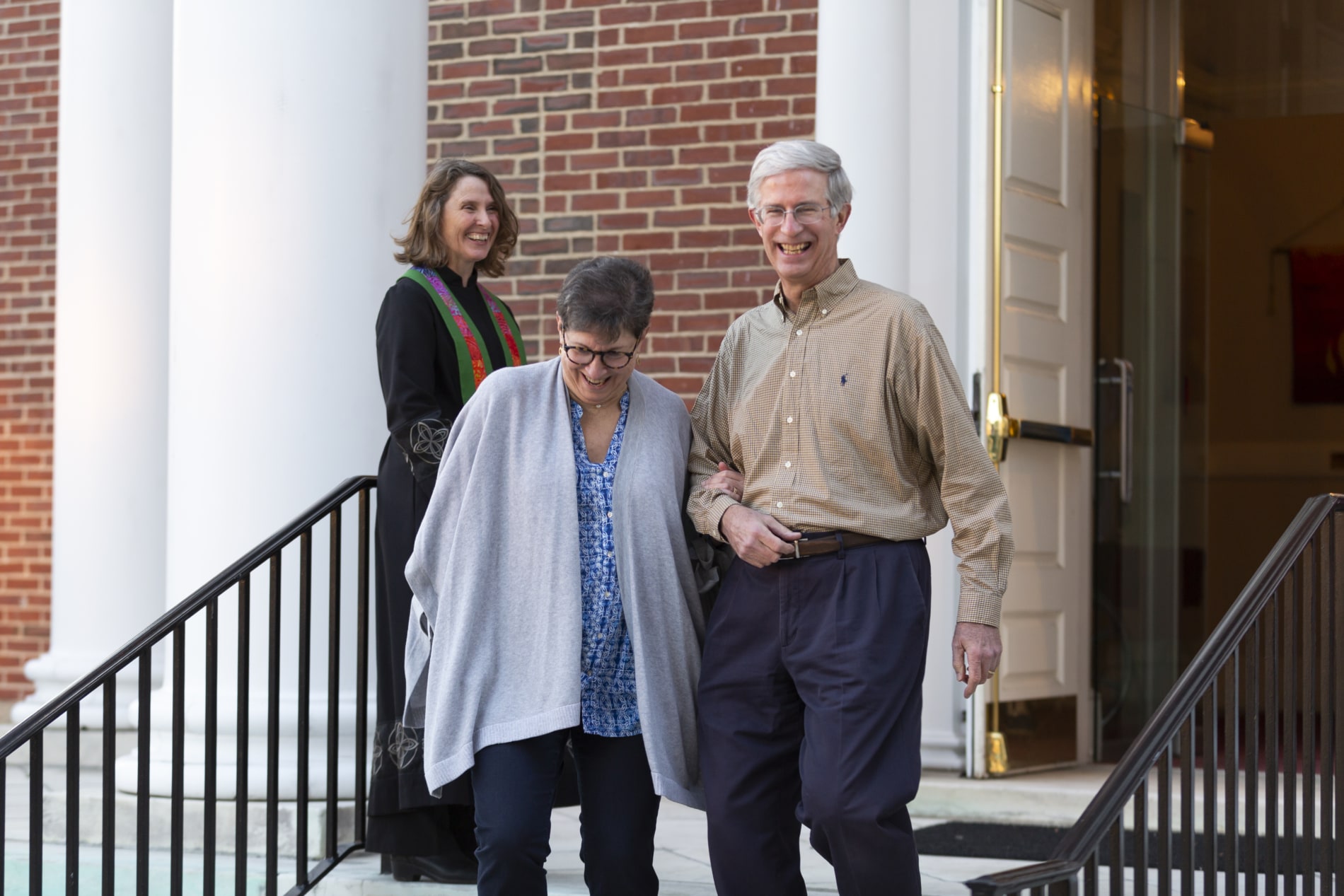 A smiling couple descends the stairs of the church while a pastor looks on