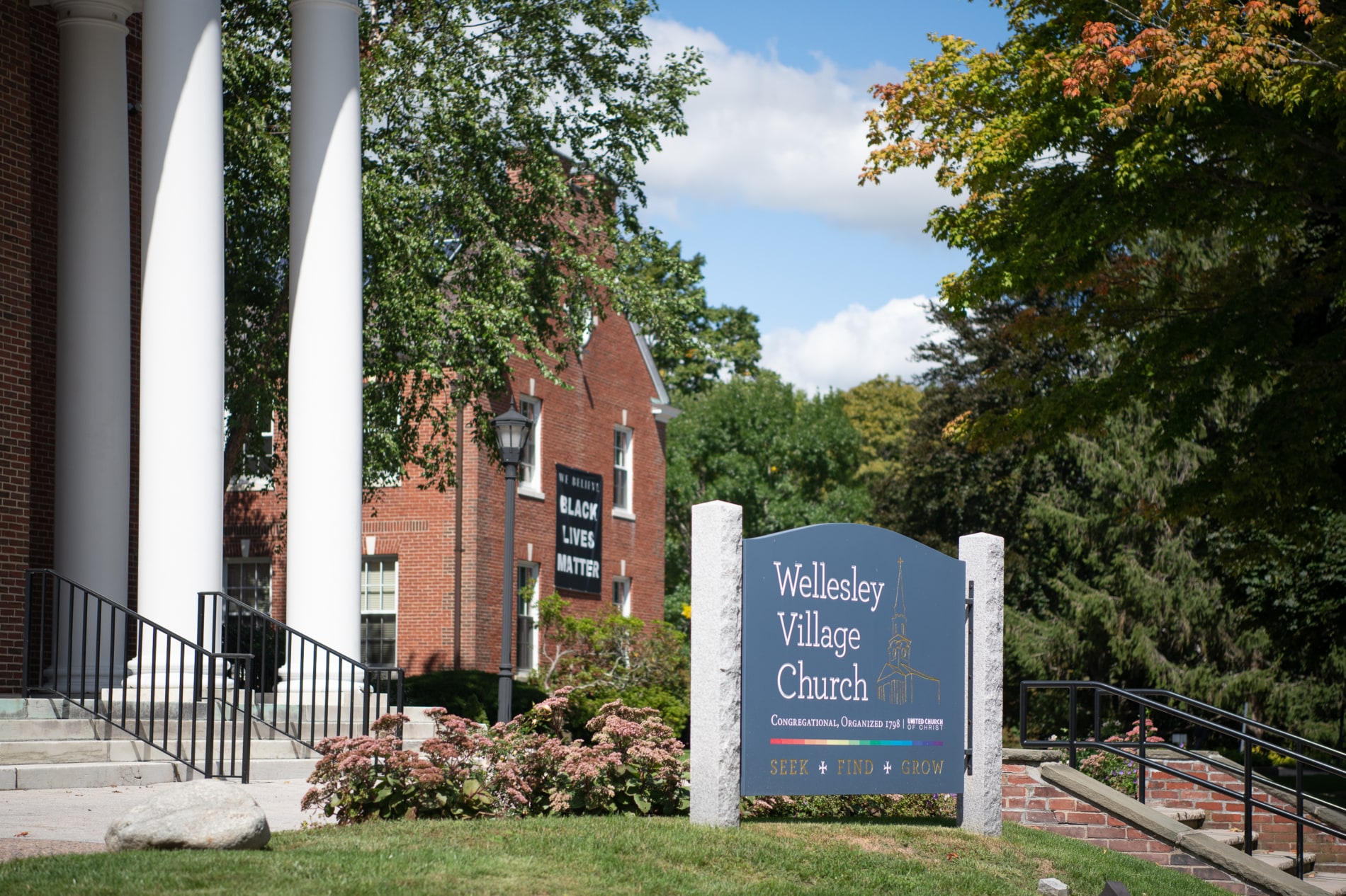 The exterior sign of the Wellesley Village Church with a Black Lives Matters banner in the background